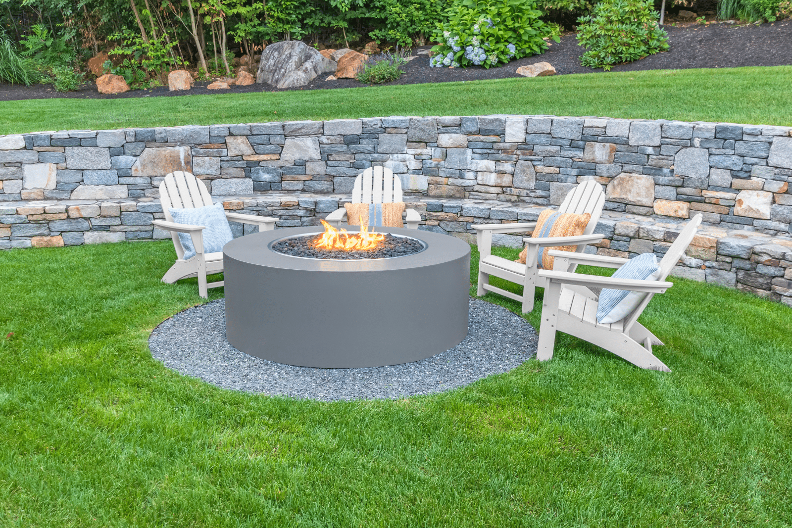 A luxury fire pit with sleek modern chairs arranged around it, set on a stone patio. The fire glows warmly, creating an inviting upscale outdoor ambiance.